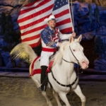 A man in a patriotic outfit rides on a white horse while holding an American flag at Dolly Parton's Stampede.