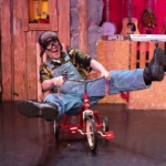 A man in goggles and overalls rides a small tricycle.