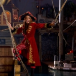A pirate captain in red hangs onto the side of a ship with weapon drawn.