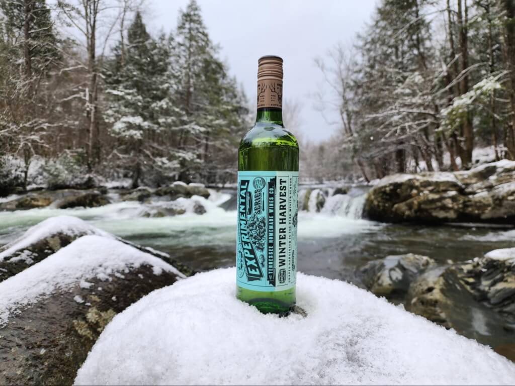 a bottle of Winter Harvest wine from Tennessee Homemade Wines sits on a snowy rock in front of a waterfall