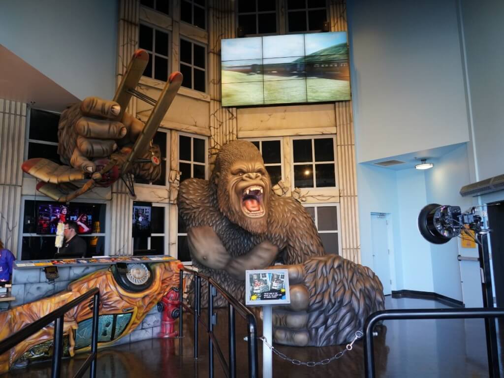 A wax gorilla statue of King Kong in a museum, standing beside a large screen displaying captivating visuals.