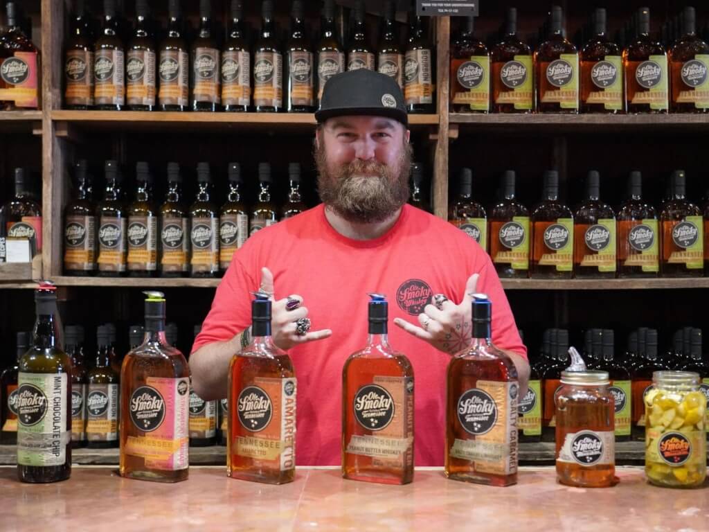 A photo of a bearded bartender standing behind a bar with moonshine bottles in front and behind him