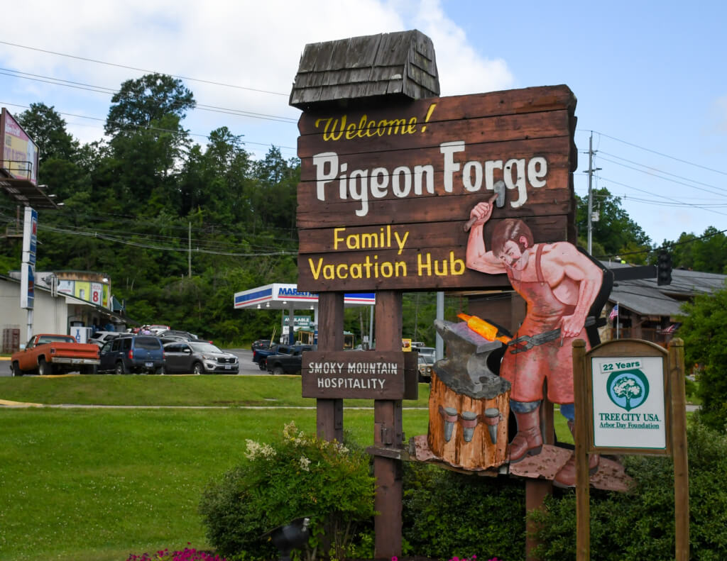 A wooden sign reading "Welcome! Pigeon Forge Family Vacation Club" next to a blacksmith working on an anvil.